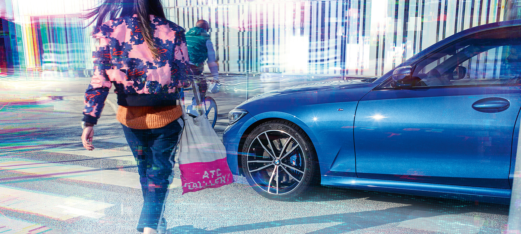 Three-quarter side shot of the BMW 3 Series Sedan on asphalt in front of an urban backround with passengers.