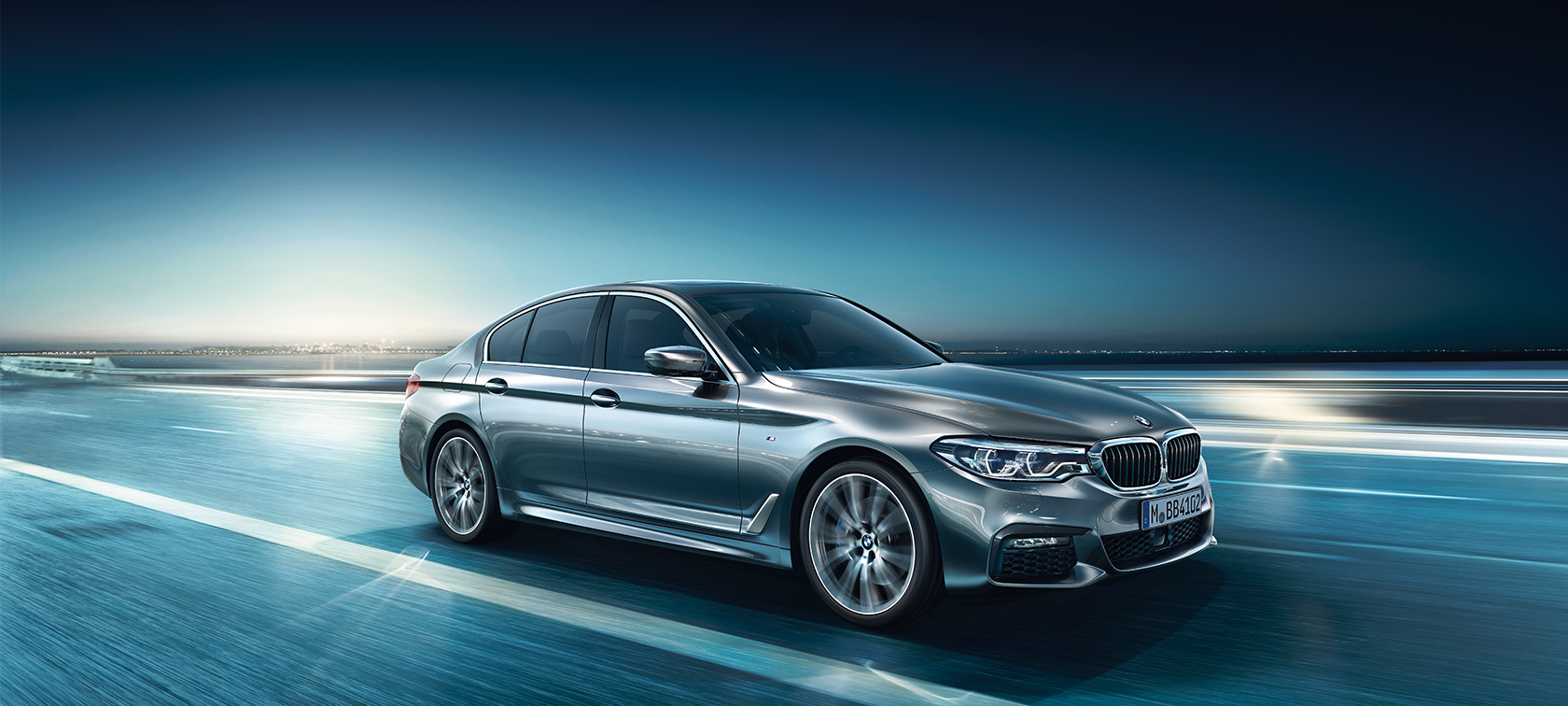 Front view of driving BMW 5 Series Sedan by day under glass building.