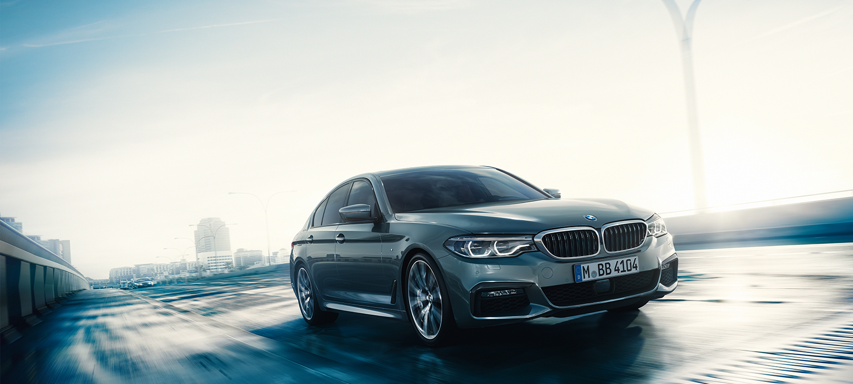 Front view of driving BMW 5 Series Sedan by day under glass building.